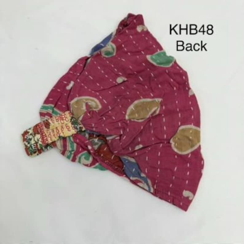 May include: A magenta fabric head wrap featuring a repeating pattern of tan and green shapes with white running stitch details. The item includes an elasticized band with a multi-colored floral print and the text KHB48 Back printed in black.