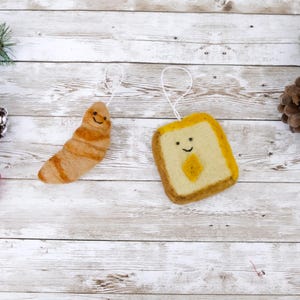 May include: Two felt food ornaments: a croissant and a slice of toast. The croissant is light brown with darker brown stripes. The toast is yellow with a brown crust and a yellow diamond in the center. Both have smiling faces and white string hangers.