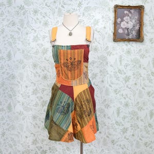 Peut inclure: Une combinaison patchwork composée de panneaux de tissu orange, vert et bordeaux à rayures verticales, agrémentée d'illustrations au trait noir représentant des papillons de nuit et des éléments botaniques, avec des bretelles et une poche poitrine.