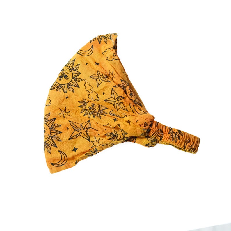 May include: A bright orange fabric headband featuring a repeating black line art pattern of suns, crescent moons, stars, and clouds. The accessory has a gathered, elasticised band section designed to provide a secure fit around the head.