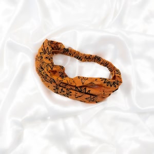 May include: An orange fabric headband featuring a repeating black line art pattern of suns, stars, and clouds. The headband has a twisted front design and an elasticised back section to allow for a secure fit around the head.