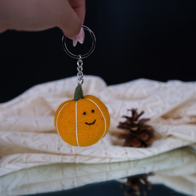 May include: A felt pumpkin keychain with a smiling face, attached to a silver key ring. The pumpkin is orange with white lines and a green stem.