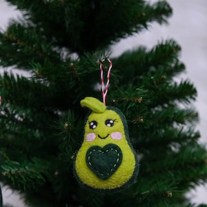 Puede incluir: Adorno navideño de aguacate hecho a mano. El adorno verde presenta un corazón verde oscuro y una cara sonriente con mejillas rosadas. Cuelga de una cuerda roja y blanca, en un árbol de Navidad.