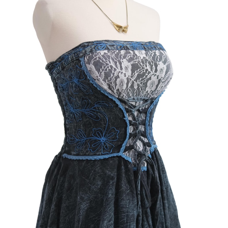 May include: A strapless dress featuring a dark acid-wash denim bodice with blue floral embroidery and a white lace panel. The bodice includes a black lace-up corset-style closure at the front, and the skirt is made of matching dark acid-wash denim.