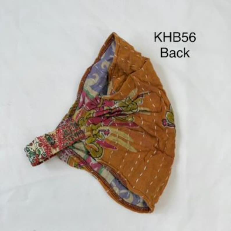 May include: A mustard yellow fabric headband featuring a floral pattern in shades of pink, green, and purple, accented with white running stitch embroidery. The item is labeled KHB56 Back and includes an elasticized section for a secure fit.