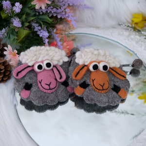 May include: Two felt sheep ornaments with white woolly heads, gray bodies, and pink and orange faces. The sheep have large black eyes and are displayed on a reflective surface. The ornaments are handmade and whimsical.