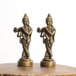 May include: Two identical brass-colored metal figurines depicting a standing figure playing a flute. Each figure wears traditional ornate attire with a textured bodice, draped lower garments, and a pointed headdress, all mounted on a circular base.
