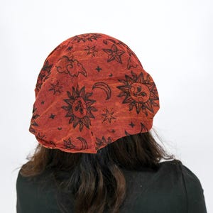May include: A rust-coloured beanie hat featuring a repeating black print of suns, crescent moons, stars, and clouds. The fabric has a textured appearance, and the design covers the entire surface of the headwear in a consistent pattern.