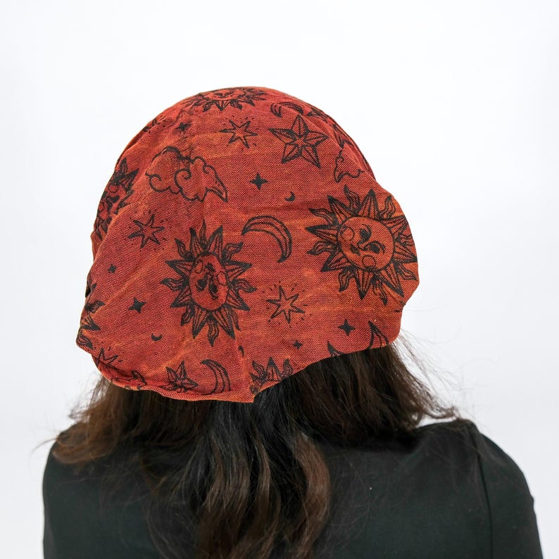 May include: A rust-coloured beanie hat featuring a repeating black print of suns, crescent moons, stars, and clouds. The fabric has a textured appearance, and the design covers the entire surface of the headwear in a consistent pattern.