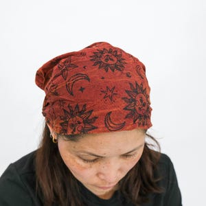 May include: A rust-coloured fabric head covering featuring a repeating black print of suns, crescent moons, and stars. The material has a textured appearance and is worn in a slouchy style, covering the top of the head with a gathered fit.