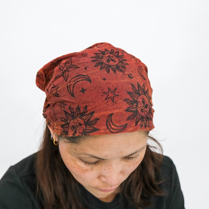 May include: A rust-coloured fabric head covering featuring a repeating black print of suns, crescent moons, and stars. The material has a textured appearance and is worn in a slouchy style, covering the top of the head with a gathered fit.