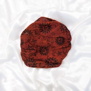 May include: A rust-coloured fabric head wrap or bonnet featuring a repeating black print of suns, crescent moons, and stars. The accessory has a gathered, circular shape with a textured surface, showcasing celestial motifs across the entire fabric.