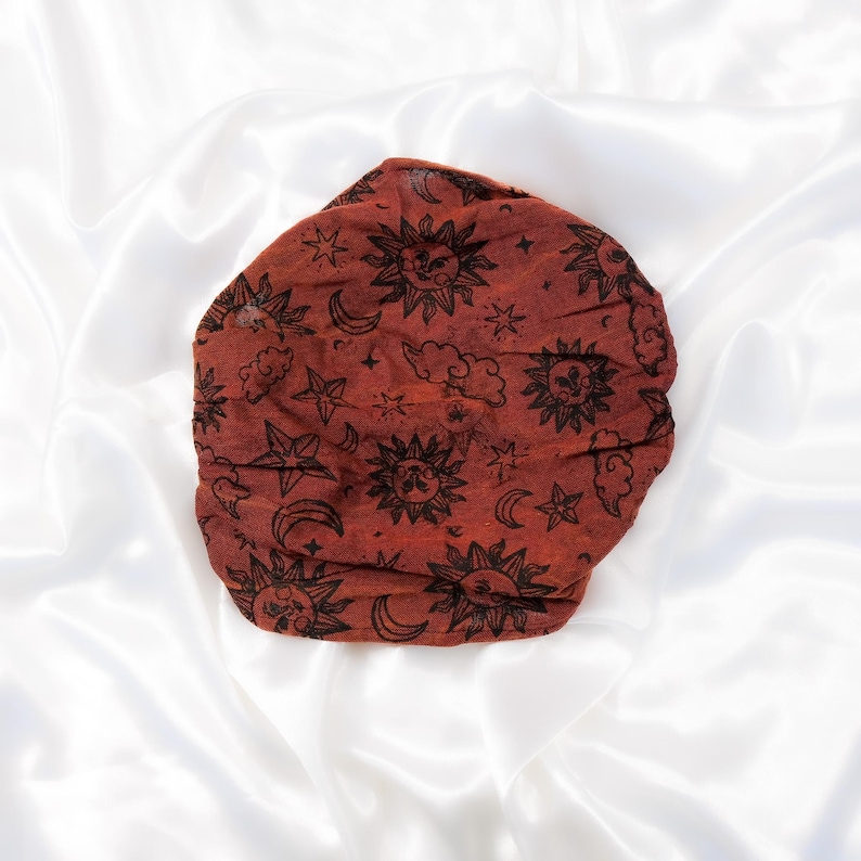 May include: A rust-coloured fabric head wrap or bonnet featuring a repeating black print of suns, crescent moons, and stars. The accessory has a gathered, circular shape with a textured surface, showcasing celestial motifs across the entire fabric.