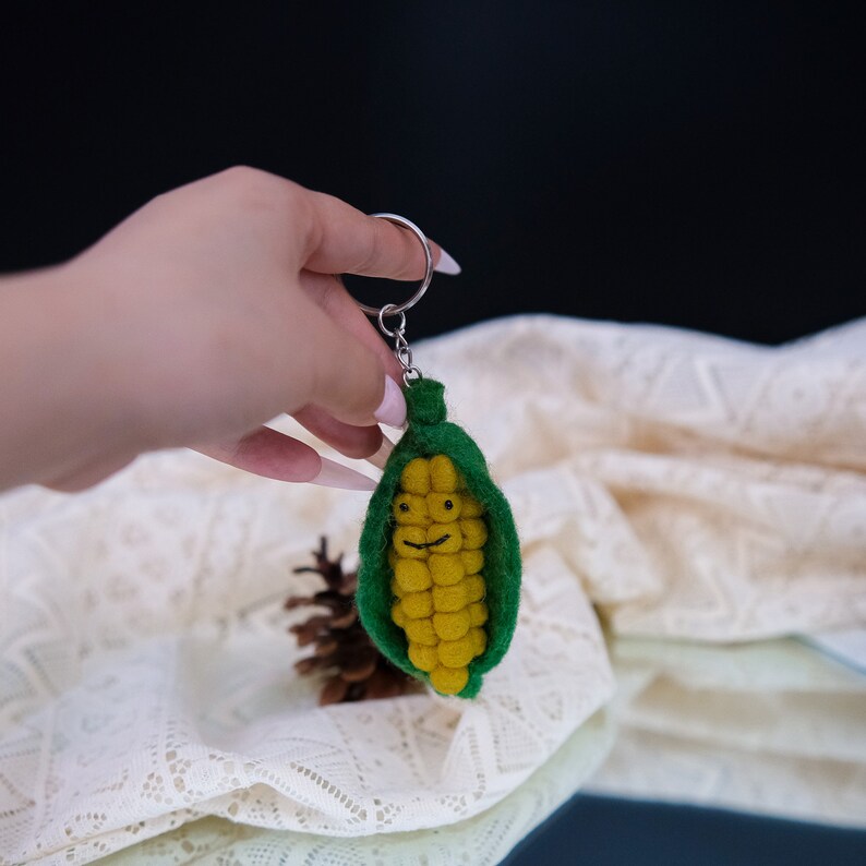 May include: A felt corn on the cob keychain with a smiling face. The corn is yellow and the husk is green. The keychain is attached to a silver ring.