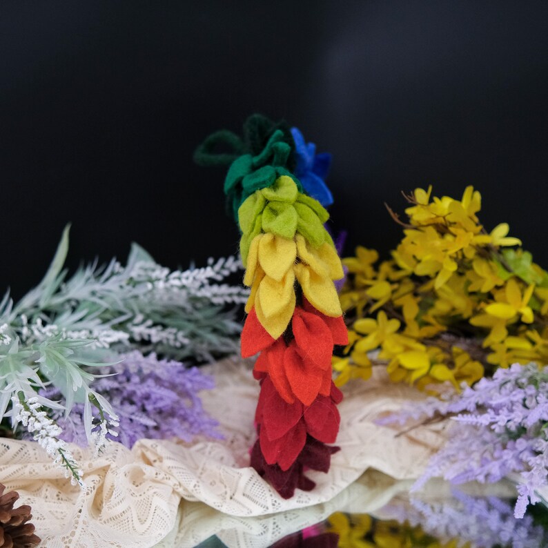 May include: A rainbow-colored felt garland with a series of leaves in shades of green, yellow, orange, and red. The garland is displayed on a white lace surface with yellow and purple flowers.