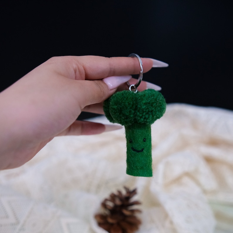 May include: A green felt broccoli keychain with a smiling face. The keychain is attached to a silver ring.