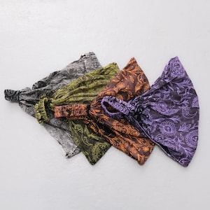 Pode incluir: Uma coleção de cinco faixas de tecido para a cabeça em várias cores, incluindo preto, verde-oliva, laranja e roxo. Cada faixa apresenta uma parte traseira elástica e um padrão floral tipo paisley. As faixas estão dispostas na diagonal.