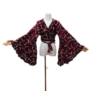 May include: A deep burgundy velvet wrap top featuring long, wide bell sleeves and a tie-front closure. The fabric is adorned with an intricate, swirling floral embroidery pattern in gold and white thread, accented with small, colorful beaded details.