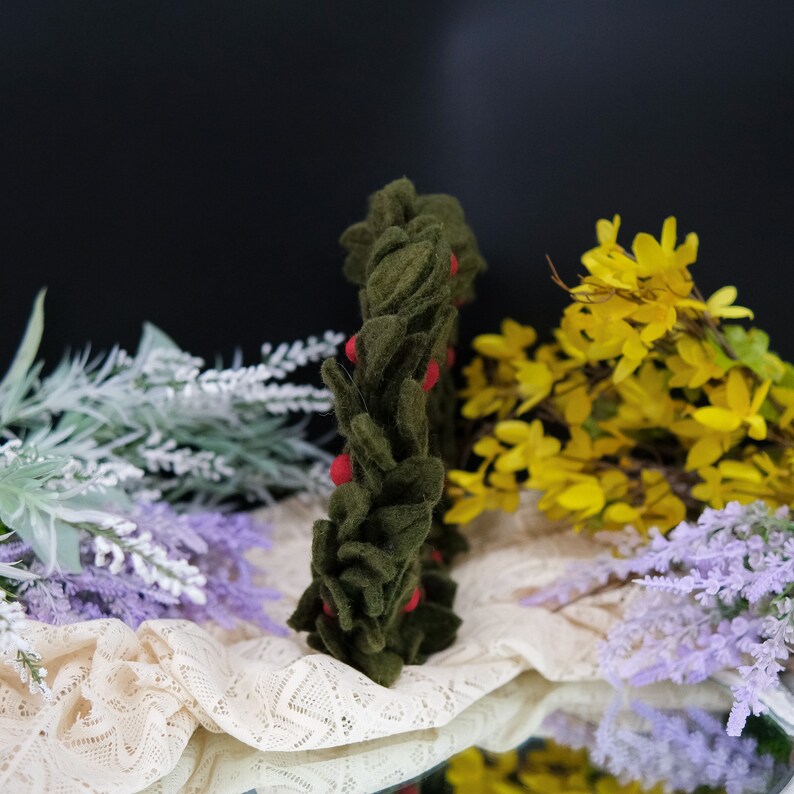 May include: A green felt wreath with red felt berries. The wreath is sitting on a white lace tablecloth with yellow and purple flowers in the background.