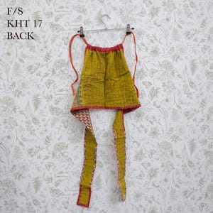 May include: A mustard yellow quilted halter top featuring a grid pattern and red trim, with long fabric ties extending from the top corners. The text F/S KHT 17 BACK is printed in the upper left corner of the image.