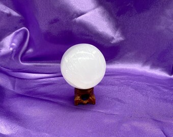 Puede incluir: Una esfera de selenita blanca sobre un soporte de madera. La esfera es lisa y pulida, con un ligero brillo. El soporte es marrón oscuro y tiene un diseño sencillo.