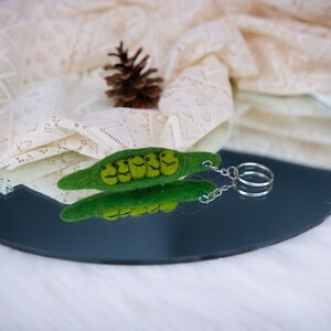 May include: A green felt pea pod keychain with five smiling peas inside. The keychain has a silver metal ring and chain.