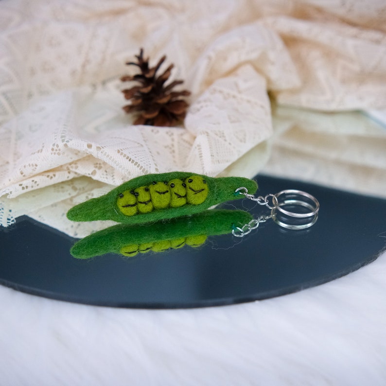 May include: A green felt pea pod keychain with five smiling peas inside. The keychain has a silver metal ring and chain.