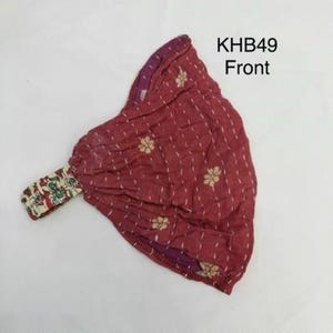 May include: A maroon fabric headband labeled KHB49 Front, featuring a repeating pattern of small white floral motifs and vertical dashed lines. The accessory includes a gathered elastic section with a multi-colored floral print on a white background.
