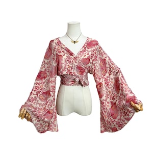 May include: A cream-colored wrap top featuring a vibrant red and pink floral and botanical print. The garment has a deep V-neckline, a tie-waist closure, and long, wide bell sleeves. A gold-toned moth pendant necklace is worn at the neck.