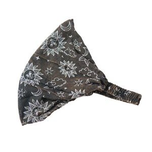 May include: A dark grey fabric headband featuring a white line-art pattern of suns, crescent moons, stars, and clouds. The accessory includes a gathered elastic band section at the back to provide a secure fit for the wearer.