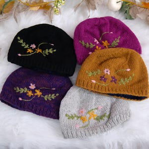 May include: Five knitted beanies in black, magenta, mustard yellow, purple, and light gray. Each beanie features a ribbed brim and an embroidered floral design with green leaves, yellow flowers, and small pink or purple blossoms.