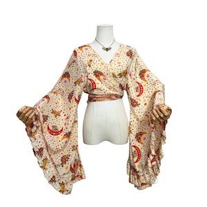 May include: A cream-colored wrap top featuring a celestial print with red and orange crescent moons, stars, and floral motifs. The garment has long, wide bell sleeves with ruffled edges and a tie-waist closure, displayed on a mannequin.