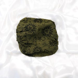 May include: An olive green fabric headwrap featuring a repeating black print of suns with faces, crescent moons, stars, and clouds. The accessory has a gathered, textured appearance and is displayed in a circular, folded configuration.