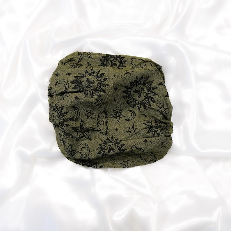 May include: An olive green fabric headwrap featuring a repeating black print of suns with faces, crescent moons, stars, and clouds. The accessory has a gathered, textured appearance and is displayed in a circular, folded configuration.