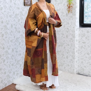 May include: A long, open-front kimono-style robe featuring a patchwork design in shades of brown, mustard yellow, and deep red. The garment is adorned with various circular and floral patterns and is worn over a white top and a long white skirt.