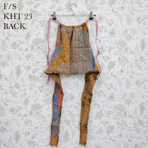 May include: A multi-colored apron featuring a patchwork design with brown, yellow, blue, and orange tones, hanging on a metal hanger. The apron has long fabric ties and visible stitching throughout. The text F/S KHT 23 BACK is printed in black.