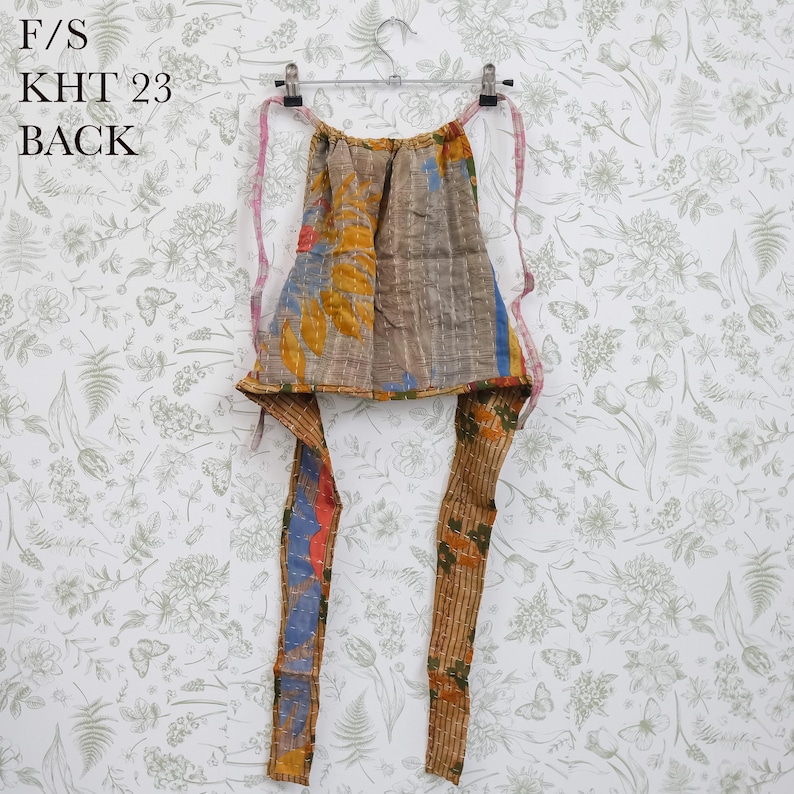 May include: A multi-colored apron featuring a patchwork design with brown, yellow, blue, and orange tones, hanging on a metal hanger. The apron has long fabric ties and visible stitching throughout. The text F/S KHT 23 BACK is printed in black.