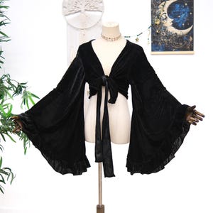 May include: A black velvet tie-front top featuring long, wide bell sleeves with ruffled cuffs. The garment has a deep V-neckline and long fabric ties at the center front that allow for an adjustable closure, displayed on a mannequin.