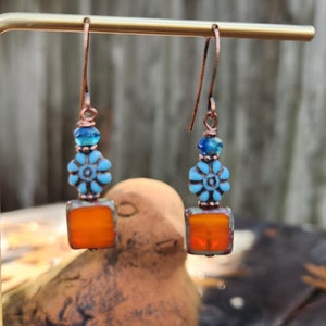 May include: A pair of dangle earrings with a copper wire design. The earrings feature a square orange glass bead, a blue flower bead, and a small blue glass bead.