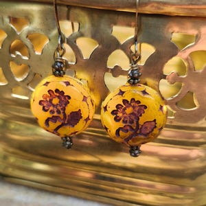 May include: A pair of handcrafted earrings featuring yellow, spherical beads with a floral design in shades of brown. The earrings have dark metal accents and are suspended from simple ear wires. The background is a gold-colored decorative container.