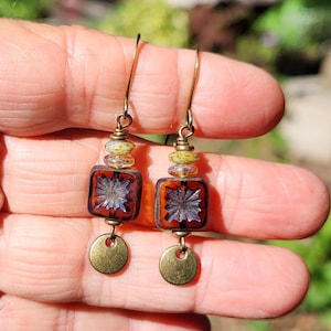 May include: Dangle earrings featuring a square, dark amber-coloured glass bead with a central starburst pattern, accented by a smaller speckled bead above and a flat, circular brass-toned metal charm at the bottom, all suspended from a gold-toned wire hook.