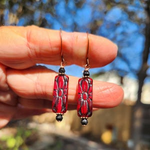 May include: A pair of red and black earrings with a floral design. The earrings have a rectangular shape with a flower pattern carved into the red glass. The earrings are suspended from copper-colored hooks and have black bead accents.