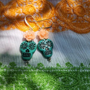 May include: A pair of turquoise skull earrings with black floral designs and peach-coloured rose accents. The earrings have silver-coloured hooks and small silver-coloured beads at the bottom. The earrings are displayed on a white and yellow lace background.