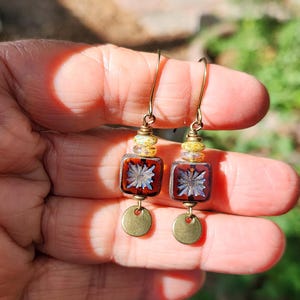 May include: A pair of dangle earrings featuring a gold-tone hook, a small yellow and white speckled bead, a square dark amber glass bead with a central starburst pattern, and a small circular brass-coloured metal disc at the bottom of each earring.
