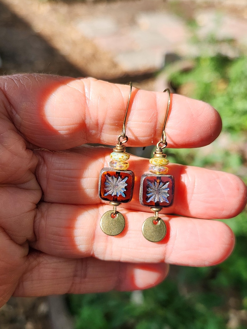 May include: A pair of dangle earrings featuring a gold-tone hook, a small yellow and white speckled bead, a square dark amber glass bead with a central starburst pattern, and a small circular brass-coloured metal disc at the bottom of each earring.