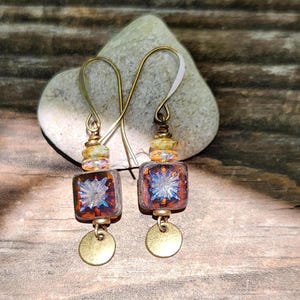 May include: A pair of drop earrings featuring square, amber-coloured glass beads with a blue starburst pattern. Each earring includes a small, round, metallic gold-toned disc at the bottom, suspended from a brass-toned hook with decorative beads.