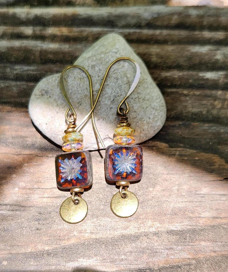 May include: A pair of drop earrings featuring square, amber-coloured glass beads with a blue starburst pattern. Each earring includes a small, round, metallic gold-toned disc at the bottom, suspended from a brass-toned hook with decorative beads.