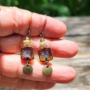 May include: A pair of dangle earrings featuring bronze-toned ear wires, small yellow and iridescent glass beads, square amber-coloured glass beads with a silver-toned starburst pattern, and small circular bronze-toned metal charms at the base.