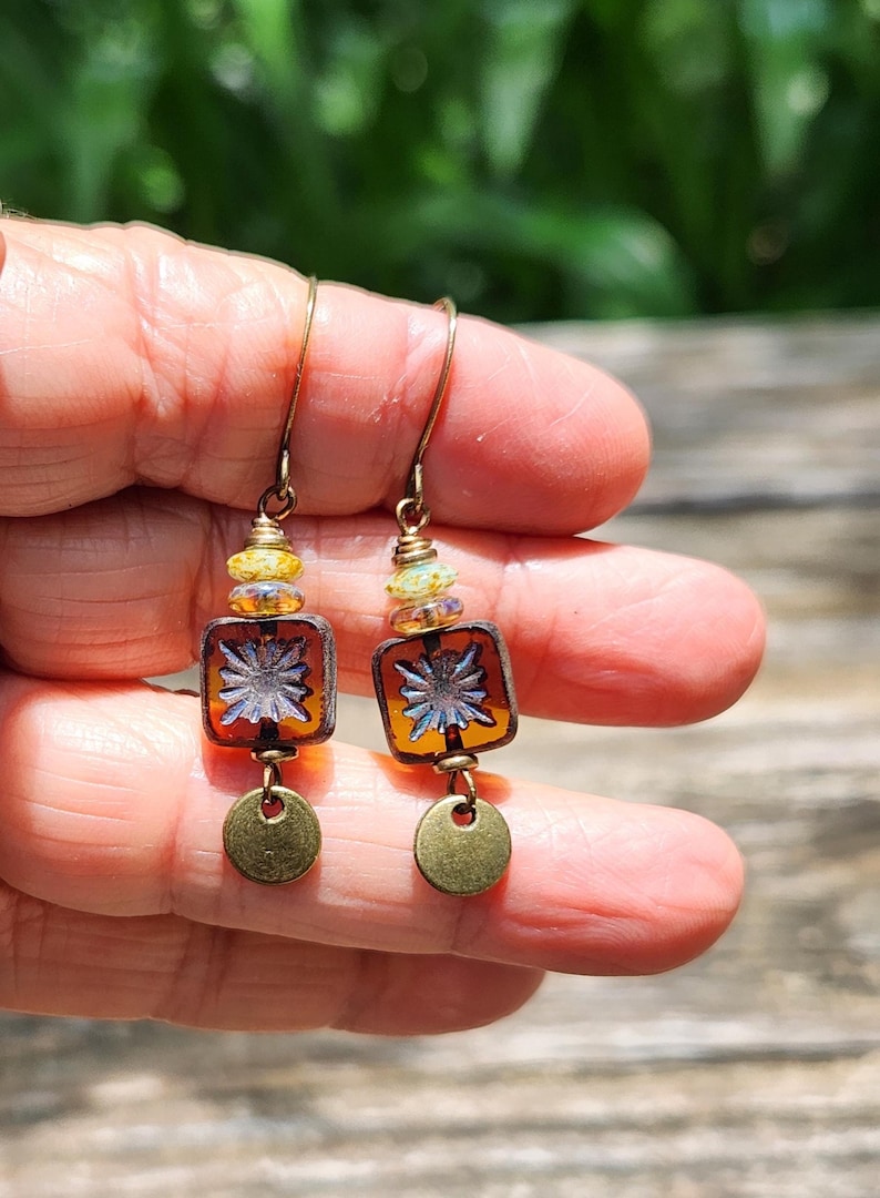 May include: A pair of dangle earrings featuring bronze-toned ear wires, small yellow and iridescent glass beads, square amber-coloured glass beads with a silver-toned starburst pattern, and small circular bronze-toned metal charms at the base.
