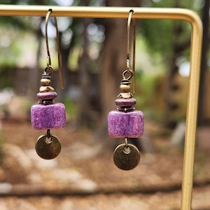 May include: A pair of dangle earrings with a gold-tone finish. Each earring features a stack of beads, a textured purple square bead, and a small, round, gold-tone charm. The earrings hang from a gold-tone ear wire.